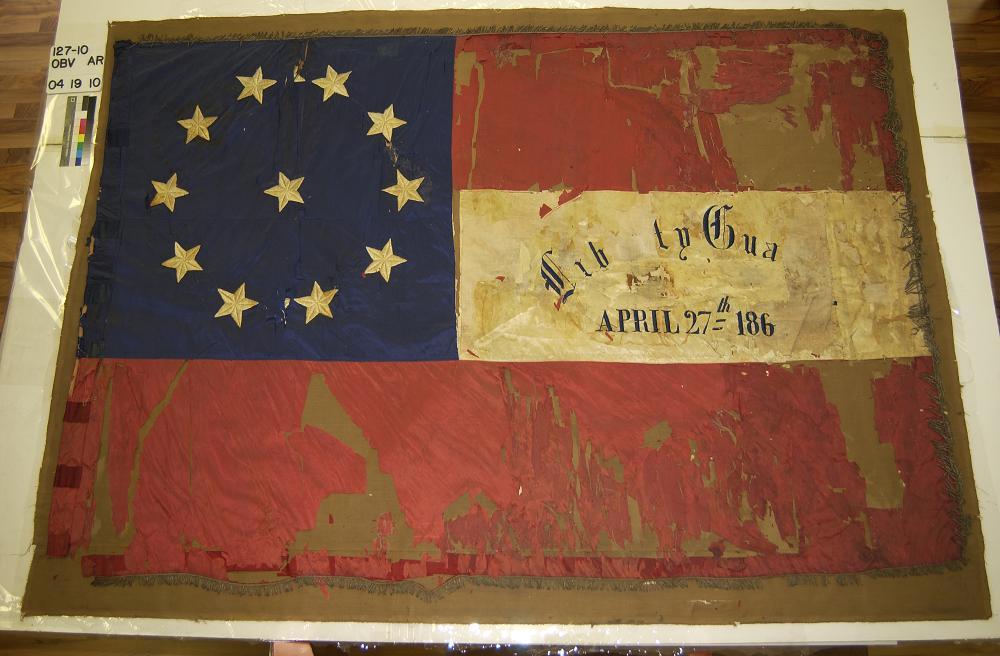 A picture of the Confederate Army battle flag of the 22nd Mississippi Infantry housed at the Mississippi Department of Archives and History in Jackson, MS. The flag is is comprised of eleven white stars in a field of blue in the upper left corner of the flag, as well as two red bars and a single white bar containing the words "Liberty Guards, April 27th 186(?)."